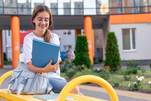 A girl with a backpack near the school and a blue notebook. On the background of the school. . High quality photo