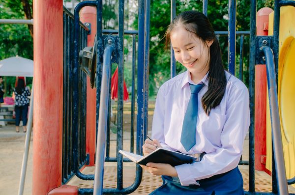 Lovely teenage girl wearing school uniform writing book while sitting on a swing at the park